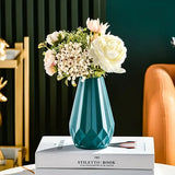 Vaso geométrico azul-petróleo com arranjo de flores claras sobre um livro